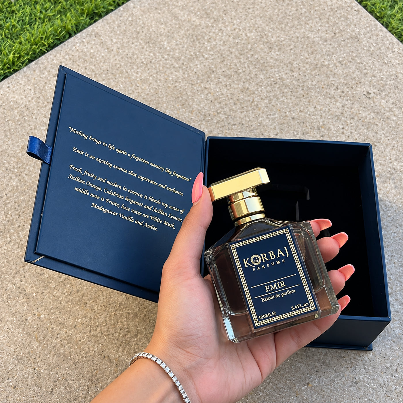 Korbaj Emir 100ml unisex citrus perfume: hand-held bottle beside an open navy box on stone.