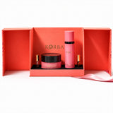 Front view of Korbaj Her Signature body gift set: pink body spray, dusting powder jar and two perfume minis in an open coral box.