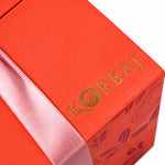 Gold Korbaj logo and pink satin ribbon on the coral Her Signature scented body gift set box, close-up detail.